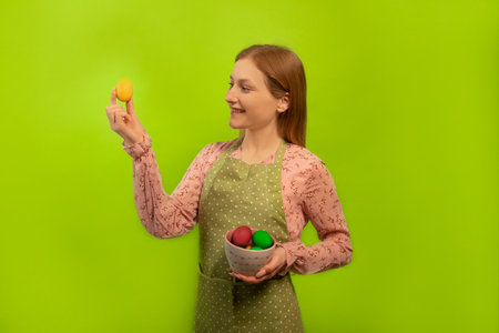 Happy smiling girl in green polka dot kitchen apron raising hand with yellow painted easter egg and keeping piala bowl with colored dyed easter eggs isolated on green background.の写真素材