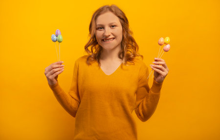 Happy smiling girl with wavy hair holding small painted dyed easter eggs on sticks, wearing knitted yellow sweater, celebrating Easter Day isolated on colorful yellow background.の写真素材