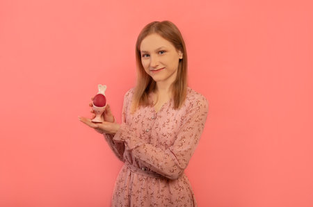 Happy cheerful smiling woman holds rabbit egg stand with one red painted egg, wears pink floral dress and looks at camera isolated on pink background. Easter or spring concept.の写真素材