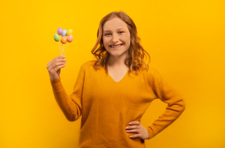 Happy smiling laughing young woman in yellow sweater holding small painted dyed easter eggs on sticks, keeping hand on waist hip isolated on yellow background.の写真素材