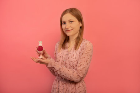 Happy cheerful smiling girl holding rabbit bunny egg stand with red painted egg, wearing pink floral dress and looking aside isolated on pink background. Easter and spring concept.の写真素材