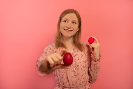 Happy smiling girl in spring pink dress holds two red painted easter eggs or shows dyed easter eggs isolated on pink background. Young woman holds out one hand with colorful egg.の写真素材