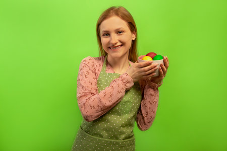 Happy smiling young woman wearing green polka dot kitchen apron and holding piala bowl cup with multicolored painted dyed easter eggs isolated on green background.の写真素材