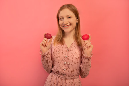 Happy smiling laughing girl holds two red painted easter eggs or shows dyed easter eggs and wears spring dress with floral pattern print isolated on pink background. Easter Day.の写真素材