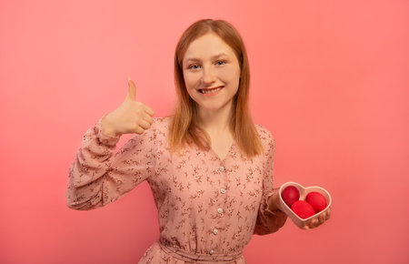 Happy smiling woman showing thumb up or keeping like gesture hand and holding heart shaped plate dish with three dyed painted easter eggs isolated on pink background. Easter Day.の写真素材