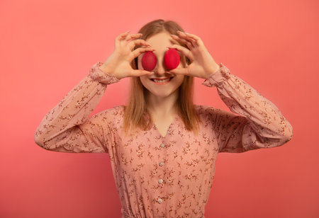 Happy smiling young woman holding two red painted easter eggs or two dyed easter eggs on eyes making joyful face expression isolated on pink background. Girl celebrating Easter.の写真素材