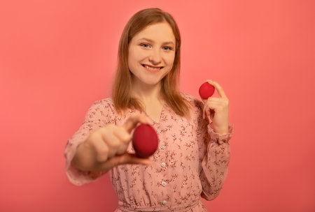 Happy smiling woman in floral pink dress holds two red painted easter eggs or shows dyed easter eggs isolated on pink background. Girl puts one hand forward with colored egg.の写真素材