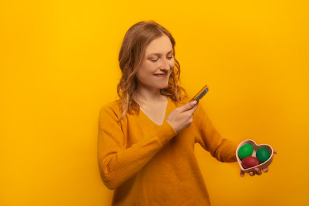 Happy smiling girl in yellow sweater holding heart shaped plate saucer with three multicolored painted easter eggs, taking photo of dyed easter eggs isolated on yellow background.の写真素材