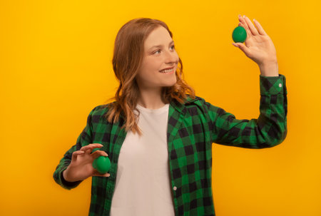 Happy smiling young woman holding two green painted dyed easter eggs, making choice or choosing, wearing green checkered plaid shirt with white shirt isolated on yellow background.の写真素材