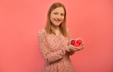 Happy smiling young woman in pink spring floral dress holds heart shaped plate with three painted easter eggs or shows dyed easter eggs isolated on pink background. Easter Day.の写真素材