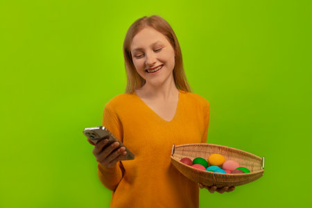 Happy smiling laughing woman in yellow sweater holding wicker basket with multicolored painted dyed easter eggs and using mobile phone or smartphone isolated on green background.の写真素材