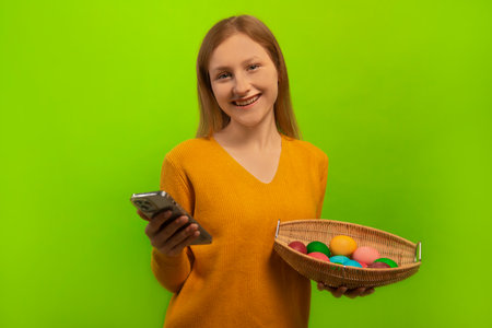 Happy smiling laughing woman in sweater holding wicker basket with multicolored painted dyed easter eggs and using mobile phone or smartphone isolated on green background.の写真素材