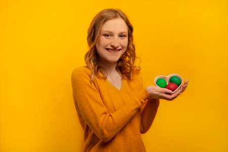 Happy smiling cheerful girl holding heart shaped plate saucer with three colorful painted dyed easter eggs, wearing yellow sweater isolated on yellow background. Easter Day.の写真素材