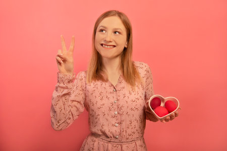 Happy smiling dreamy girl showing peace gesture v sign with two fingers hand and holding heart shaped plate dish with three dyed painted easter eggs isolated on pink background.の写真素材