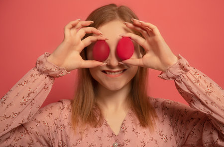 Happy smiling girl holding two red painted easter eggs or two red dyed easter eggs near eyes making joyful face expression isolated on pink background. Woman celebrating Easter.の写真素材