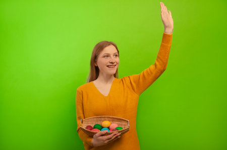 Happy cheerful smiling young woman greeting or waving hi hello, wearing sweater and holding wicker basket with colorful painted dyed easter eggs isolated on green background.の写真素材