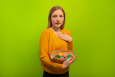 Open eyed surprised shocked girl holds wicker basket with colorful painted dyed easter eggs, wears yellow sweater and looking at camera isolated on green background. Easter Day.の写真素材
