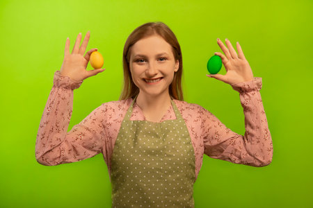 Happy smiling girl housewife in green kitchen apron with polka dots and pink dress holding two multicolored painted dyed easter eggs isolated on green background. Easter concept.の写真素材
