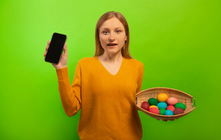 Surprised shocked girl holding wicker basket with multicolored painted dyed easter eggs and showing empty mobile phone black screen mock up isolated on green background. Easter.の写真素材