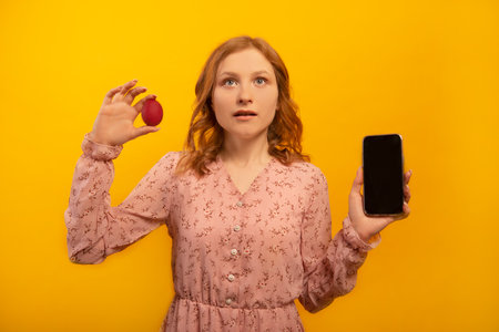 Surprised astonished shocked girl in pink dress holds red painted dyed easter egg and shows empty smartphone black screen mockup isolated on yellow background. Easter day concept.の写真素材