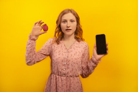 Surprised shocked young woman in pink dress holding red painted dyed easter egg and showing empty mobile phone black screen mock up isolated on yellow background. Easter concept.の写真素材