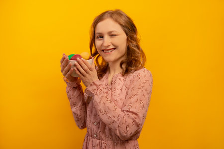 Happy smiling winking young woman wearing floral pink dress and holding piala bowl with multicolored dyed painted easter eggs isolated on yellow background. Easter day concept.の写真素材