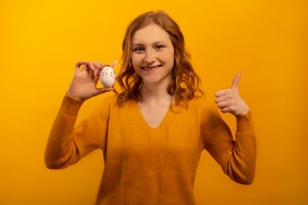 Happy smiling girl holds white decorated egg with pink rabbit ears, flowers and bunny muzzle and shows thumb up or keeps like gesture isolated on yellow background. Easter concept.の写真素材