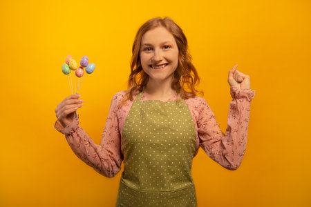 Happy smiling girl in green kitchen apron with polka dots holds multicolored painted dyed easter eggs sticks and shows winner gesture hand or keeps clenched fist on yellow background.の写真素材