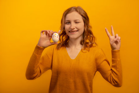 Happy smiling winking girl holding white egg decorated with pink rabbit ears, flowers and bunny muzzle and showing peace v sign gesture isolated on yellow background. Easter concept.の写真素材