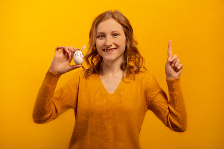 Happy smiling young woman shows white decorated egg with pink rabbit ears, flowers and bunny muzzle, points index finger up isolated on yellow background. Easter and diy concept.の写真素材