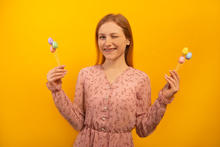 Happy smiling winking girl in pink flower dress showing multicolored painted dyed easter eggs on sticks in two hands isolated on yellow background. Easter day concept.の写真素材