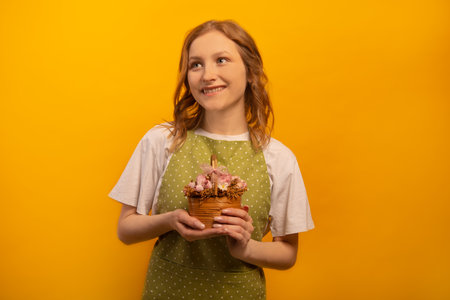 Happy smiling dreamy girl in green polka dot apron and white shirt holds pink flowers in wicker basket and looks up isolated on yellow background. Floristry floristic business concept.の写真素材