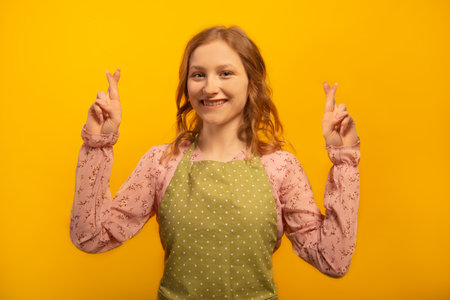 Happy smiling young woman in green polka dot kitchen apron hold fingers crossed for good luck, await for miracle, pray and hope dreams come true isolated on yellow background.の写真素材