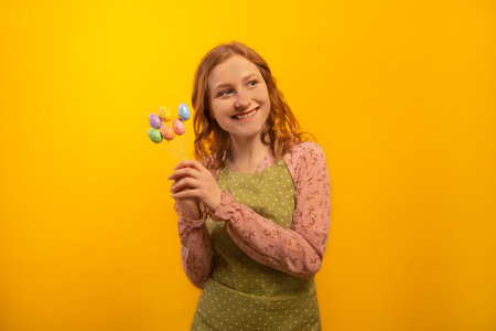 Happy smiling housewife girl in green polka dot kitchen apron holding multicolored painted dyed easter eggs sticks and looking aside isolated on yellow background. Easter day concept.の写真素材