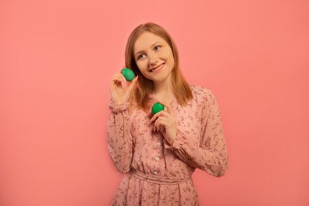 Happy smiling dreamy young woman in floral pink dress holding two green painted dyed easter eggs and looking up isolated on colored pink background. Easter day celebration concept.の写真素材