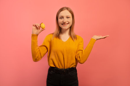 Happy smiling girl showing yellow painted dyed easter egg and holding palm up hand to insert an ad, text words isolated on pink background. copyspace. Easter day concept.の写真素材