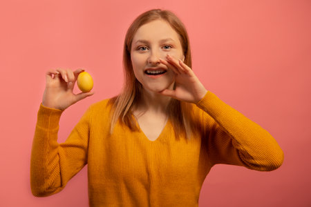 Cheerful pretty young woman in sweater holding yellow painted dyed easter egg, whispering, shouting or screaming needed information to you isolated on pink background. Easter concept.の写真素材