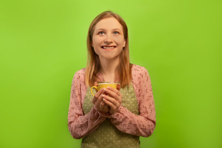 Happy smiling dreamy young woman in green polka dot kitchen apron holding yellow cup isolated on green background. Housewife resting, having break after cleaning house or cooking.の写真素材