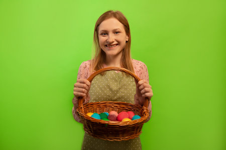 Happy smiling laughing young woman in green polka dot kitchen apron holding wicker basket with multicolored painted dyed easter eggs isolated on green background. Easter concept.の写真素材