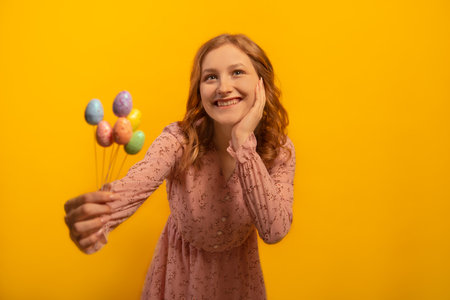 Happy smiling girl in dress showing or stretching hand to camera with multicolored painted dyed easter eggs sticks and holding hand on cheek face isolated on yellow background.の写真素材