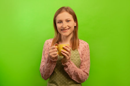 Happy smiling girl in green polka dot kitchen apron holding yellow cup isolated on green background. Housewife resting, having break after cleaning house or cooking food.の写真素材