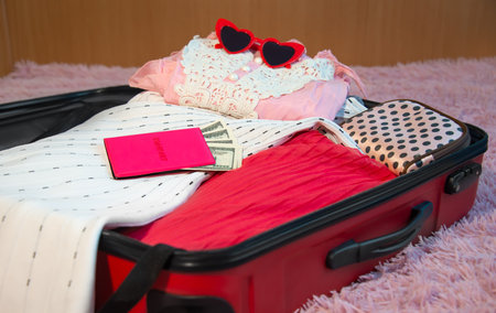 Open red suitcase of clothes, summer accessories with female pink passport with dollar bills banknotes and red heart shaped sunglasses at bed in bedroom. Travel luggage packing.の写真素材