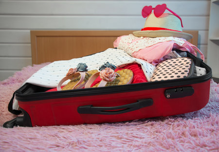 Preparation for summer holidays vacations: open red suitcase with floral slippers, female beach clothes, straw hat, cosmetic bag and sunglasses on bed. Packing things for the journey.の写真素材