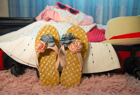 Preparing for summer holidays vacations: open red suitcase with flower beach slippers shoes, female clothes, straw hat and sunglasses on bed at bedroom. Packing things for the journey.の写真素材