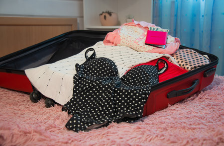 Open red suitcase full of female beach clothes, pink passport with dollar money bills, pink makeup cosmetic bag with black polka dots and black swimsuit with white polka dots.の写真素材