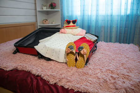 Packing things for summer holidays vacations: open red suitcase with flower beach slippers, female clothes, straw hat, red heart shaped sunglasses and pink cosmetic bag on bed.の写真素材