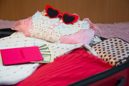 Open red suitcase of clothes, summer accessories with female pink passport with dollars bills banknotes inside and red heart shaped sunglasses on bed in bedroom. Packing luggage.の写真素材