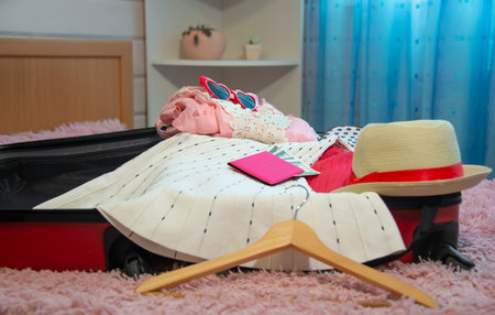 Open red suitcase full of female clothes, pink passport with dollar bills banknotes, straw beach hat, clothes hanger or clothes rack and red heart shaped sunglasses on bed.の写真素材