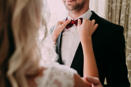 Bride fixes red bow tie on grooms neck while they stand near the windowの写真素材