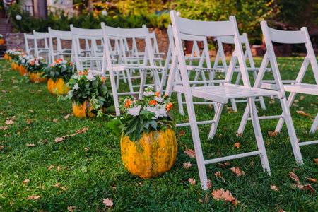 Decorating a wedding with autumn pumpkin and flowers. Ceremony outdoor in the park. White chairs for guests. Close-upの写真素材